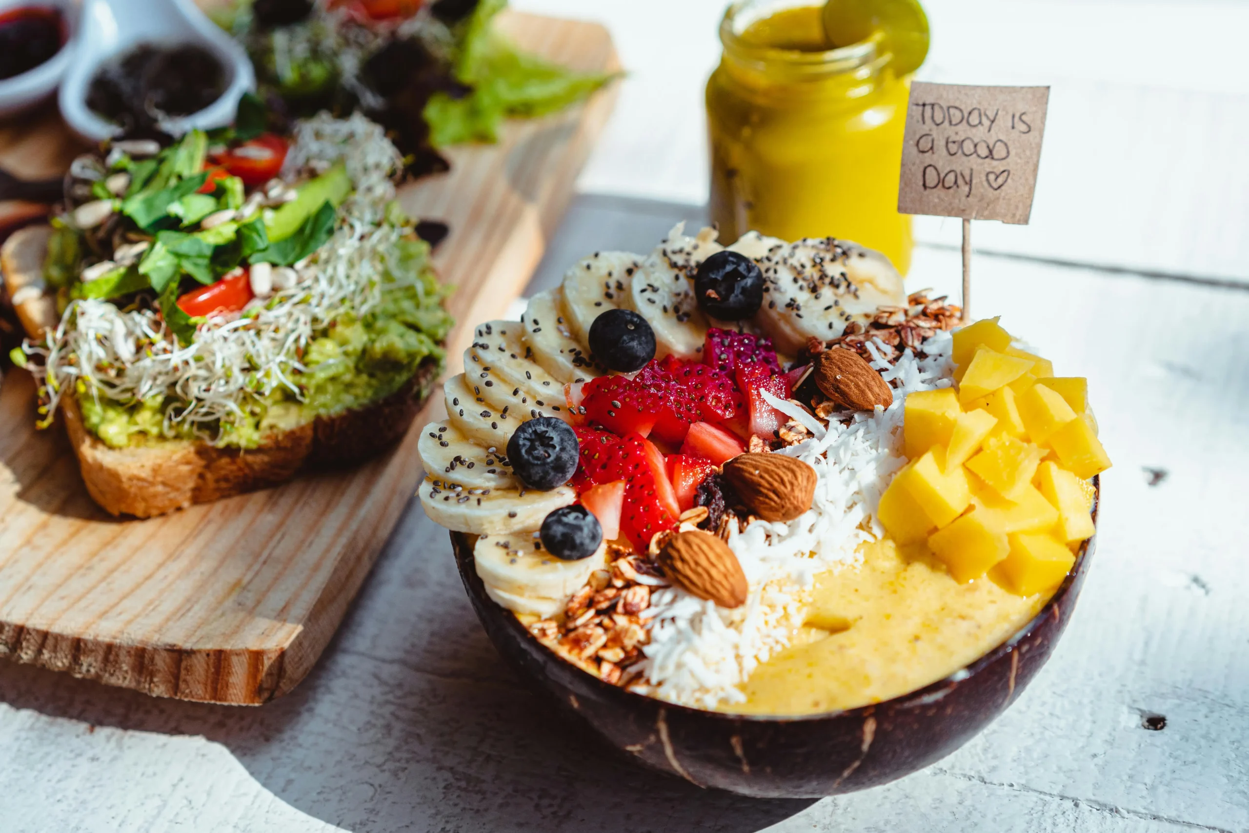A vibrant smoothie bowl topped with sliced bananas, strawberries, blueberries, mango chunks, almonds, shredded coconut, granola, and chia seeds, with a side of avocado toast on whole-grain bread. A small sign reading 'Today is a good day' is placed in the bowl, creating a positive and refreshing breakfast scene.